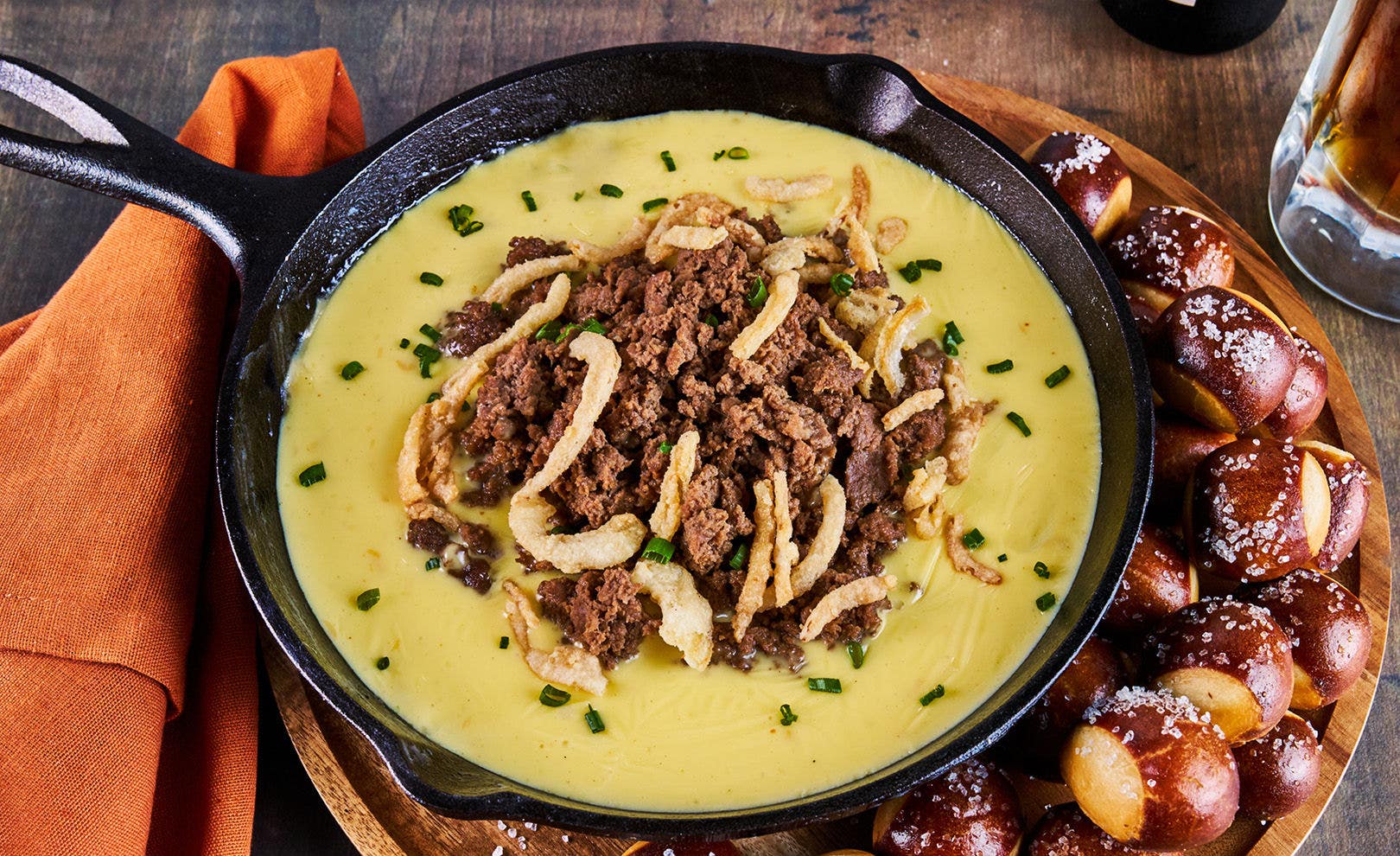 A cheese fondue in a black skillet layered with sliced beef, roasted garlic, fried shallots and chives