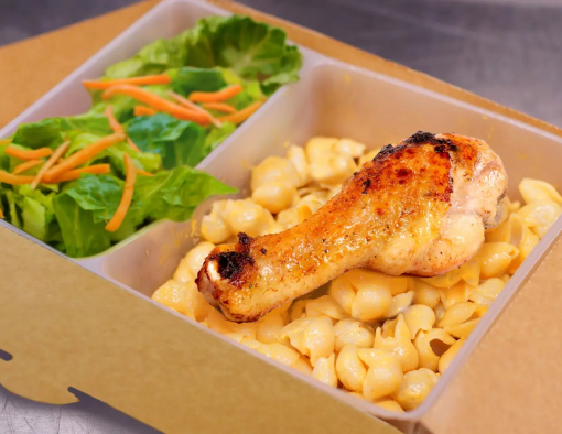 To-go box with oven-roasted drumstick on a bed of macaroni and cheese with side of lettuce and grated carrots 