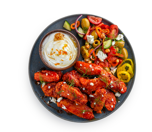 Overhead view of sauced chicken wings on a dark plate with a creamy dipping sauce, fresh salad with tomatoes and cucumbers, olives, and pickled peppers