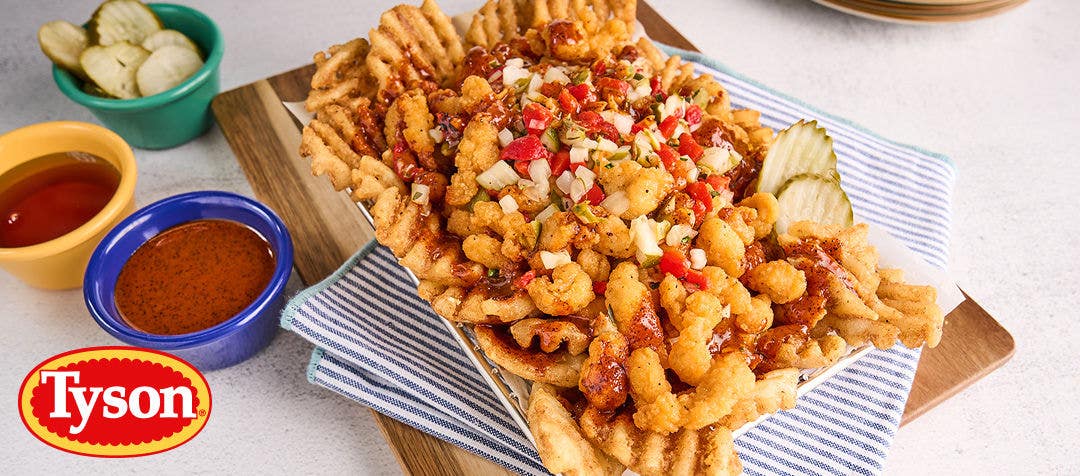 A basket of breaded mini chicken pieces on a bed of waffle fries in a basket served on a cutting board with a side of sauces and sliced pickles  