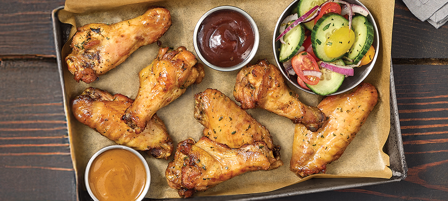 Tray of pitmaster-smoked chicken wings arranged on parchment paper with barbecue and dipping sauces, plus a cucumber tomato salad on a wooden table