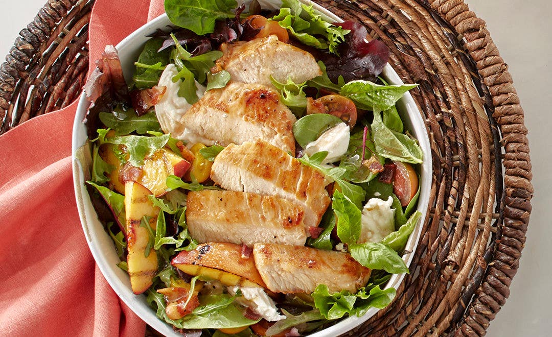 roasted chicken breast, grilled peaches, burrata cheese, and cherry tomatoes on a bed of mixed greens in a white bowl