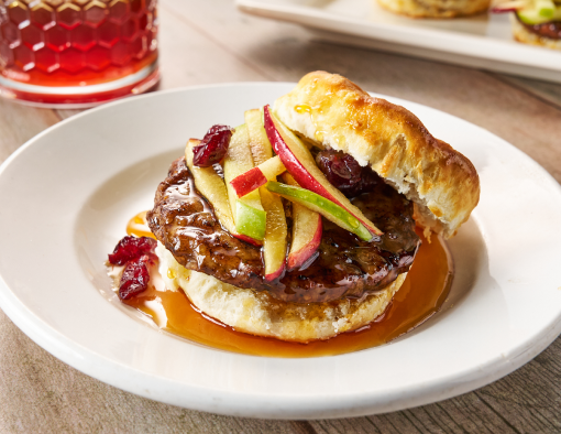 A white plate holding a pork sausage patty on a biscuit covered in syrup and topped with julienned apples and cranberries