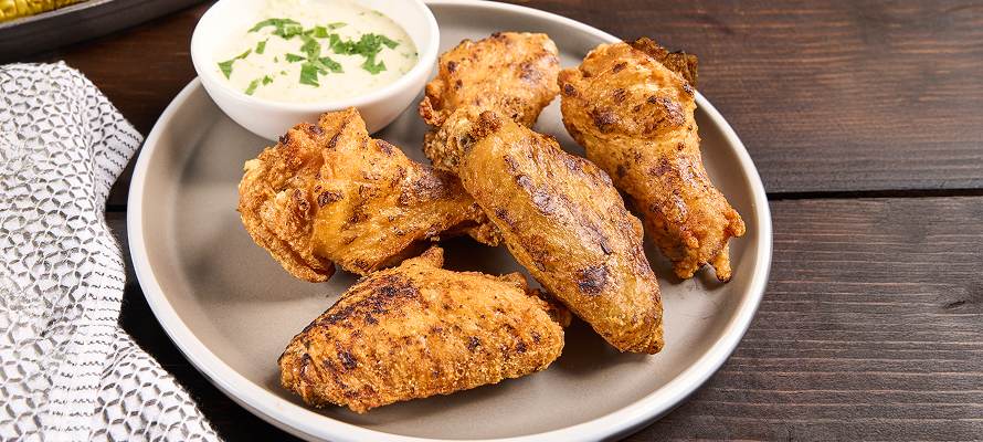 Plate of flame‑grilled chicken wings with crisp, seasoned skin, served with a creamy dipping sauce on a rustic wooden table