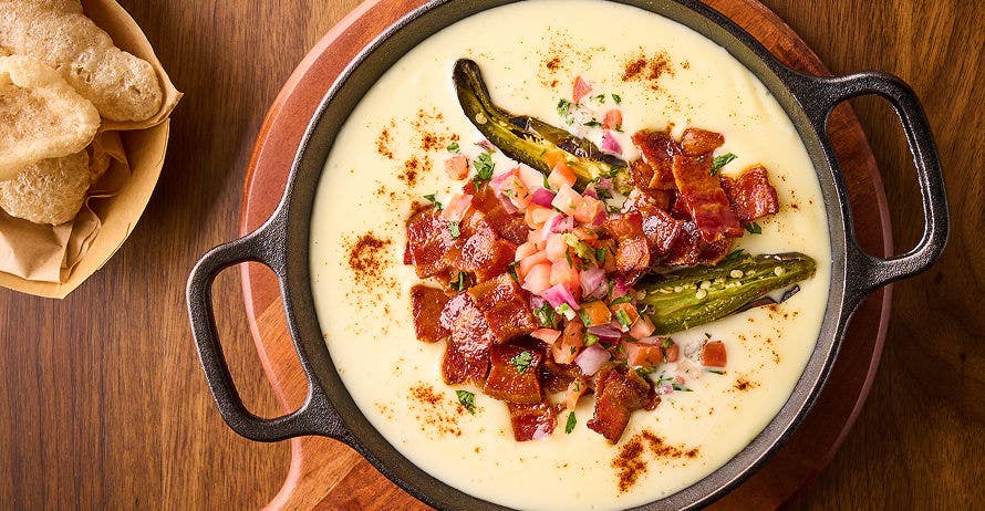 Skillet of creamy queso topped with bacon, pico de gallo, jalapeño, and chicharrones on the side