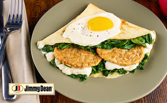 A plate with two sausage patties, a white sauce and spinach folded in a large crepé with a sunny-side-egg on top 