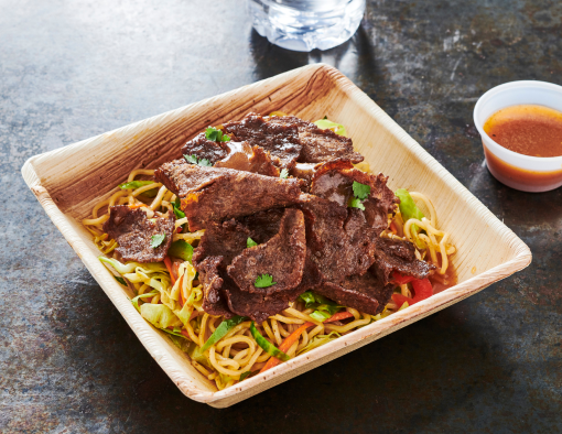 Philly beef slices over bed of noodles salad with crisp vegetables with small ramekin of dressing 