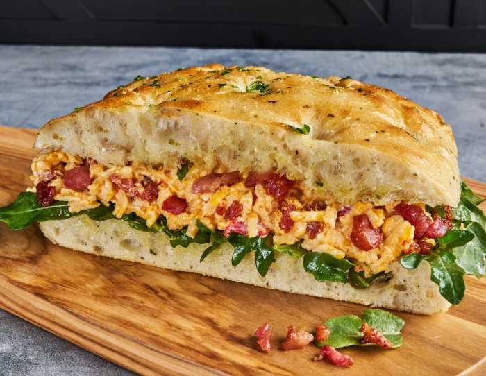 A bacon pimento cheese sandwich on focaccia bread resting on a wooden cutting board 