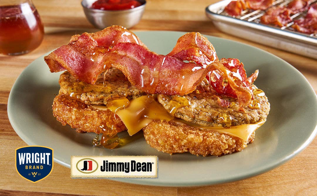 A plate with two hashbrown cakes topped with cheese, sausage patties, bacon slices and a drizzle of honey 