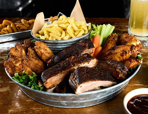 A large metal platter of barbecue with ribs, chicken wings, fries, celery, and carrots