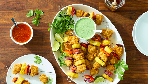 Overhead view of fried chicken skewers with pineapple, red onion, lime wedges, cilantro, and green dipping sauce on a wooden table
