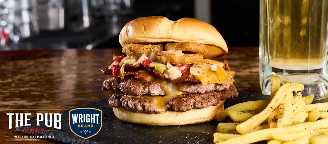 A large burger with two patties, bacon, cheese, pico de gallo, and fried pickles resting on a table with fries in the foreground  