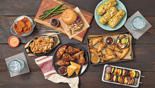 Overhead view of a table spread with chicken wings, sliders, skewers, corn on the cob, fries, sauces, and drinks on a rustic surface