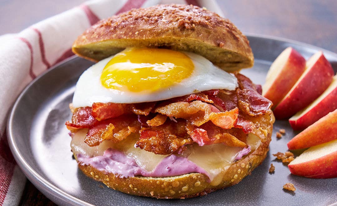 A plate with apple wedges and a cinnamon bagel sandwich with cheese, bacon and a sunny-side-up egg on top 