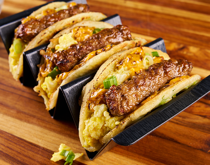 A taco holder with three pancake tacos filled with sausage links, scrambled eggs and chives  