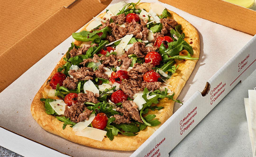 open pizza box with a flatbread pizza topped with sliced beef, cherry tomatoes, arugula and Parmesan cheese