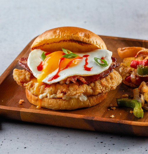Fried chicken sandwich on brioche with bacon, runny egg, sriracha, scallions on wooden plate