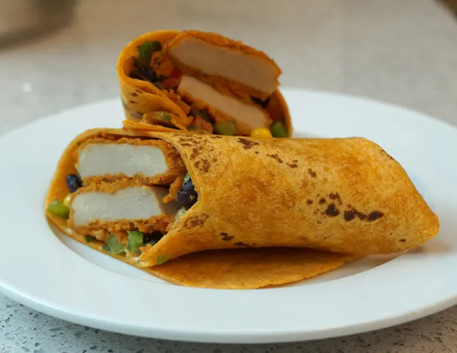 Cut breaded chicken filet, cheese sauce, black beans and corn salsa wrapped in a whole grain tortilla cut in half on a plate 