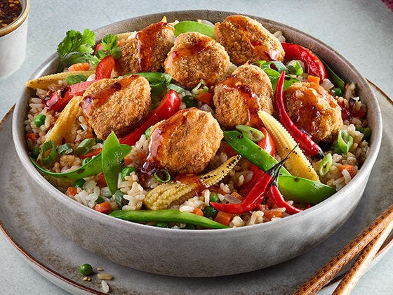 A bowl of fried rice topped with  spicy breaded beef bites, Asian style vegetables and peppers, green onion and a teriyaki sriracha sauce. 