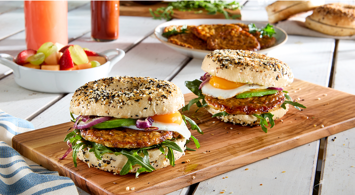Two breakfast sandwiches on everything bagels with sausage, egg, and avocado on a wooden board