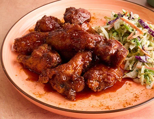 Saucy chicken wings and coleslaw on a peach plate with a small bowl of creamy dipping sauce