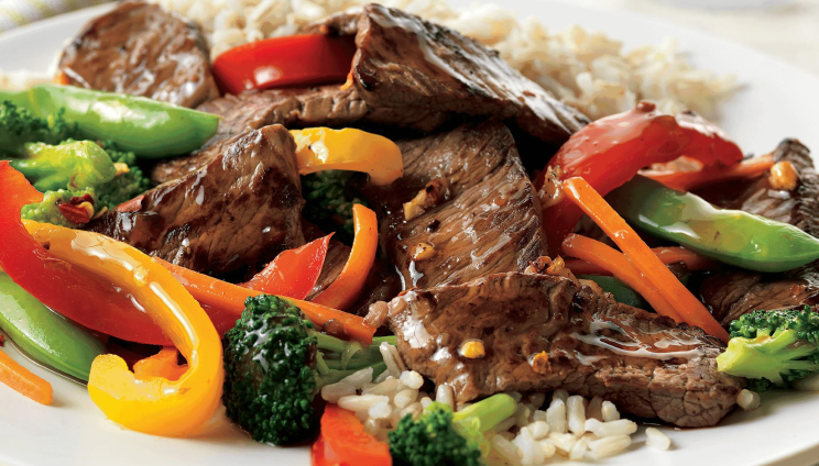 Beef stir-fry with bell peppers, broccoli, snow peas, and carrots served over rice on a white plate