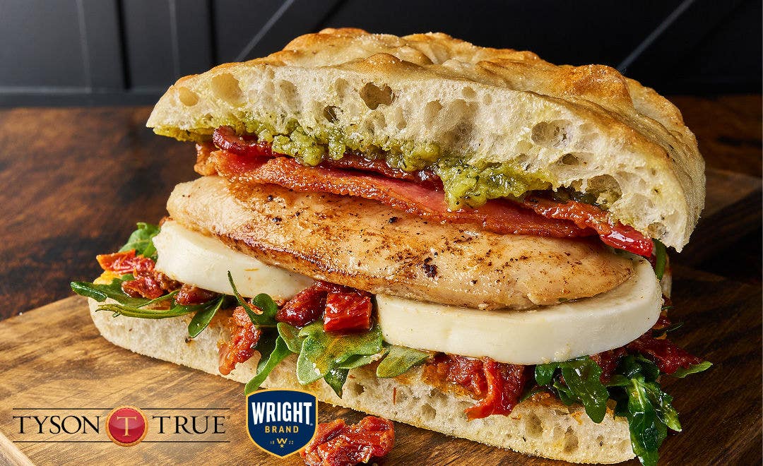 A wood tabletop with a juicy grilled chicken breast on focaccia bread with thick cuts of mozzarella cheese, sliced bacon and tomatoes. 