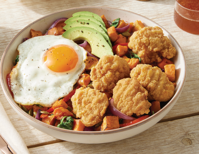 Bowl of crispy breaded chicken pieces with a fried egg, sliced avocado, roasted sweet potato hash, red onion, and greens on a table