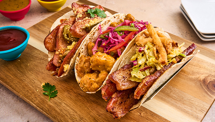 three different tacos on a wooden plate with a side of sauce