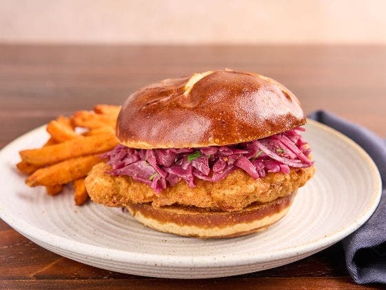 Crispy chicken schnitzel on a pretzel bun with mustard-braised red cabbage, sweet potato fries on a ceramic plate, set on a wooden table with a dark napkin