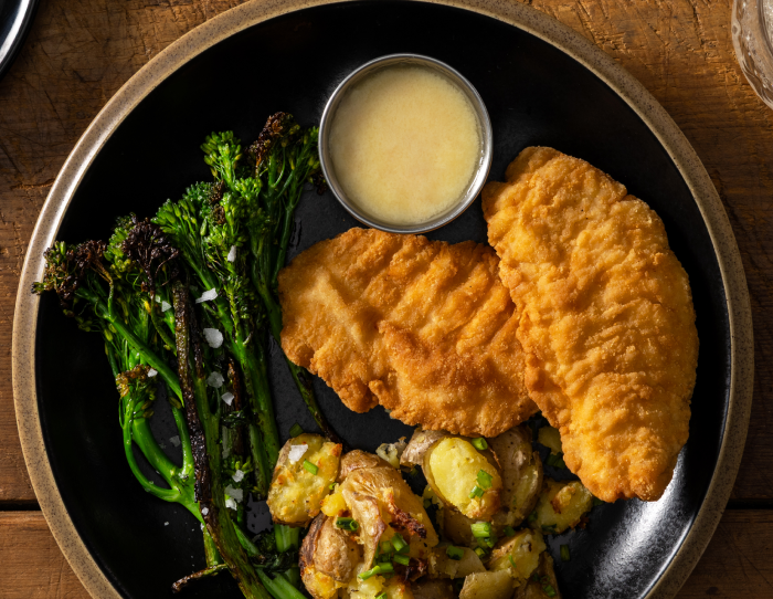 Plate with two breaded steakhouse chicken tenderloins served alongside roasted potatoes, broccolini, and a small cup of sauce