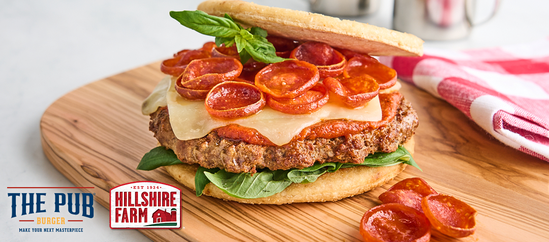 A cutting board with a burger between two small pizza crusts with cheese, marinara sauce, pepperoni and basil leaves     