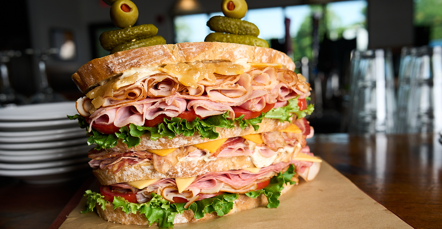 A tall, multi-layered sandwich with meats, cheeses, lettuce, tomato, with olives and pickles on skewers