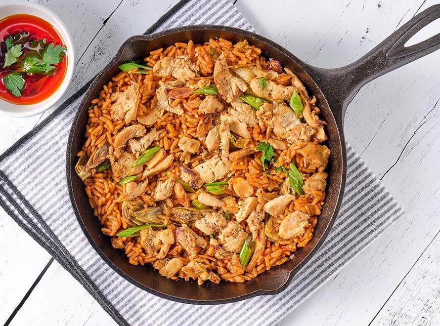 A cast-iron skillet of sliced chicken, artichoke hearts, peas and rice 