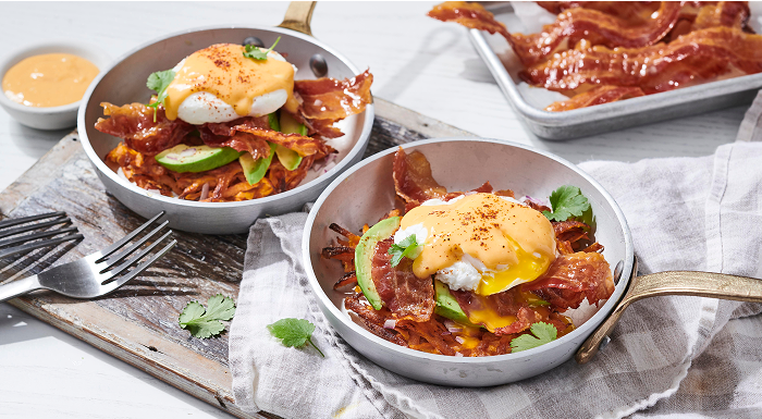 Two individual pans of eggs Benedict with bacon, avocado, and sweet potato hash