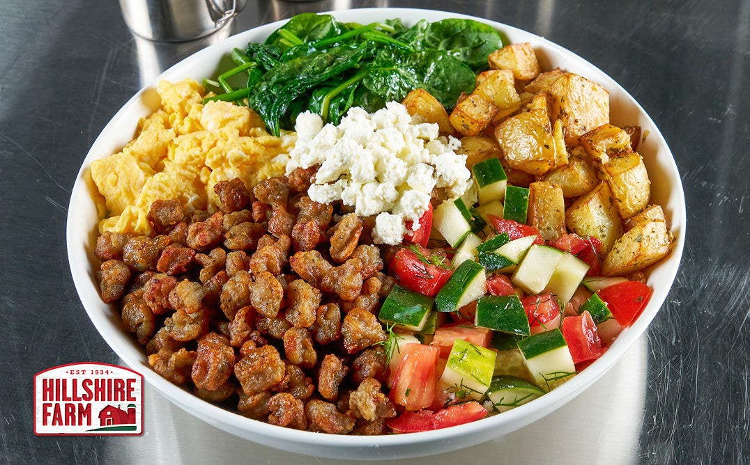 A bowl filled with sectioned-off scrambled eggs, spinach, roasted diced potatoes, sausage crumbles, cucumbers, tomatoes and feta cheese 
