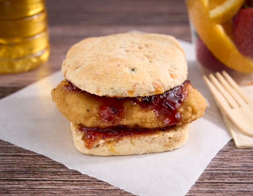 A napkin with a small, breaded chicken filet on a biscuit with jam and side of fruit 
