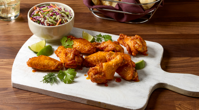 Saucy chicken wings on a board with a bowl of coleslaw, lime, and cilantro