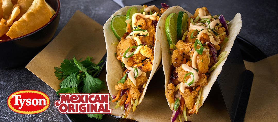 A plate with two breaded chicken tacos topped with slaw and a drizzle of sauce with a side of wonton chips          