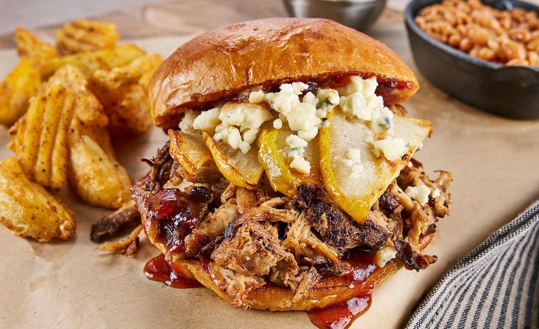 A pork barbecue sandwich stuffed with cooked pear slices and blue cheese crumbles with sides in the background