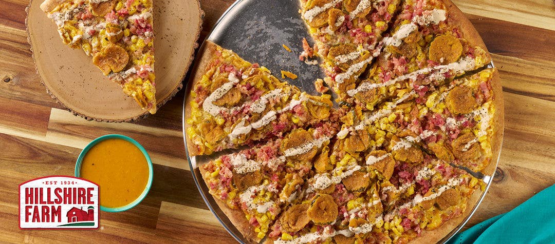 a pizza with cheese, bacon fried pickles with a side of barbecue sauce with one slice setting to the side 
