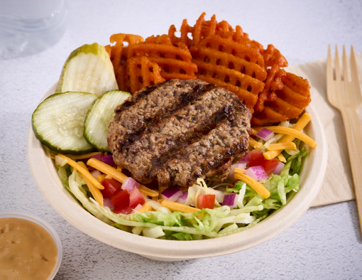A bowl with a beef patty on shredded lettuce, onions, tomatoes, sliced pickles and sweet potato fries topped with sauce