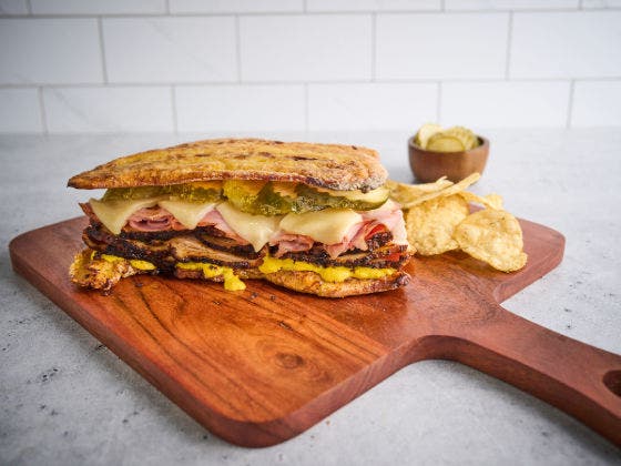 A pressed Cuban sandwich on a wooden board with chips and pickles
