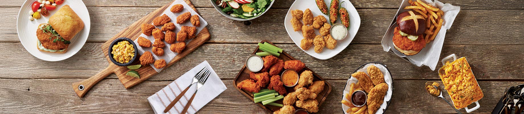 assorted fried chicken dishes on rustic wooden table, including wings, tenders, sandwiches, fries, mac and cheese, salads