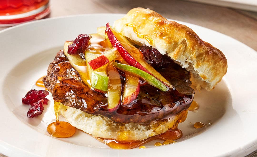 A white plate holding a pork sausage patty on a biscuit covered in syrup and topped with julienned apples and cranberries