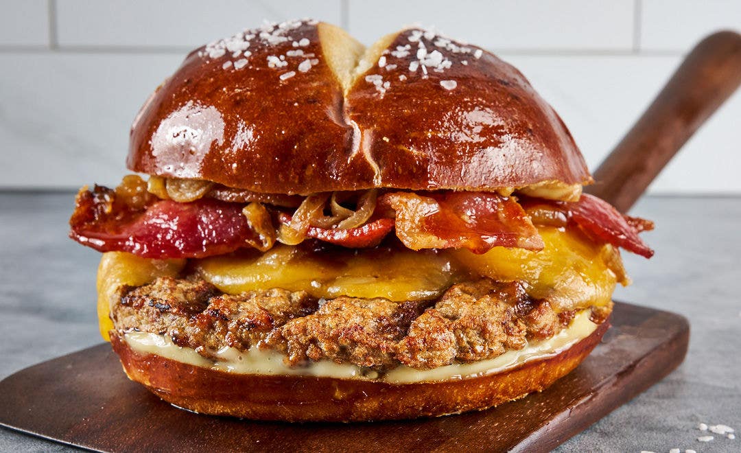 A fully loaded burger with bacon, cheese and caramelized onions spilling out of a pretzel bun 