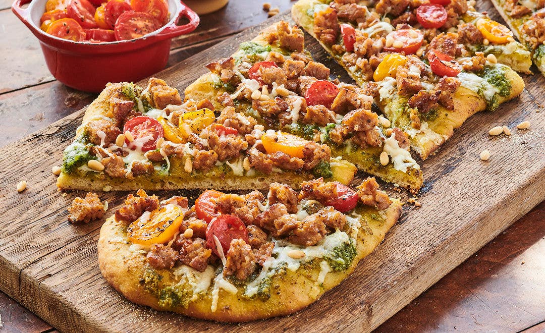 cutting board with a flatbread pizza loaded with crumbled sausage, cherry tomatoes and pesto
