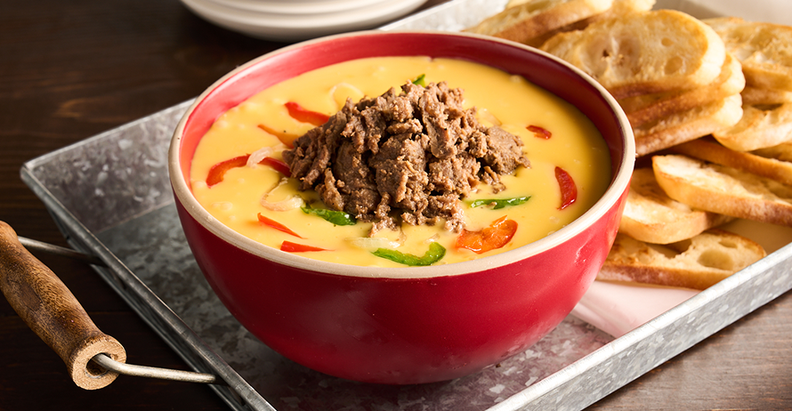 A bowl of creamy cheese dip topped with steak and peppers, served with toasted bread slices on a tray
