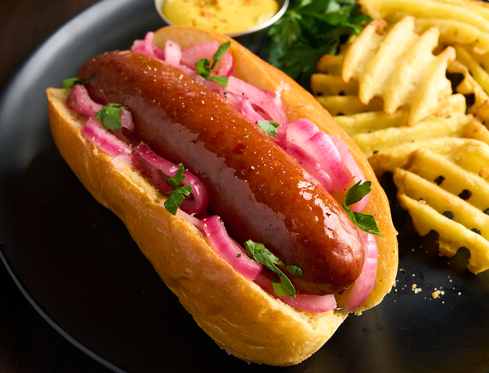 Smoked sausage in a toasted bun topped with pickled red onions and herbs, served with waffle fries and a small cup of mustard sauce