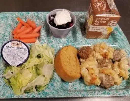 School tray of beef meatballs mixed in alfredo pasta with sides of bread, lettuce, carrots and milk